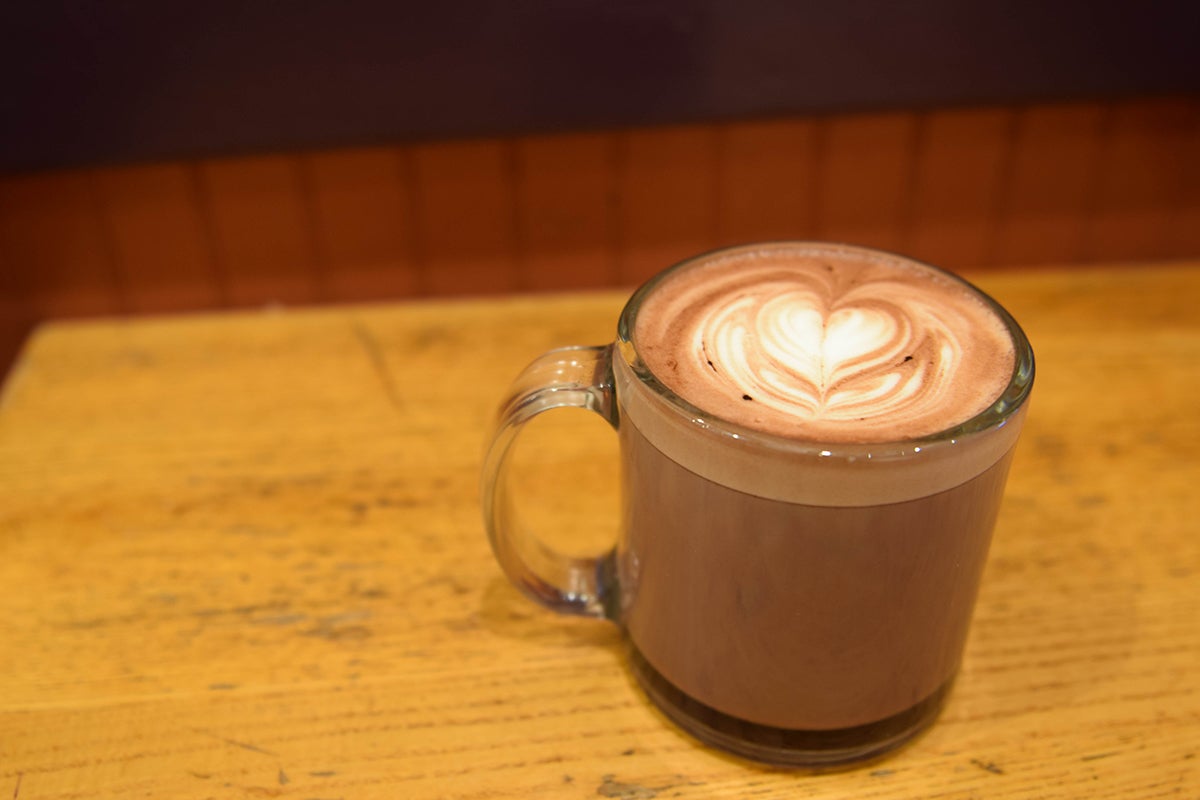 A clear glass mug of hot chocolate, decorated with a heart in the foam at the top, sits on a wooden table.