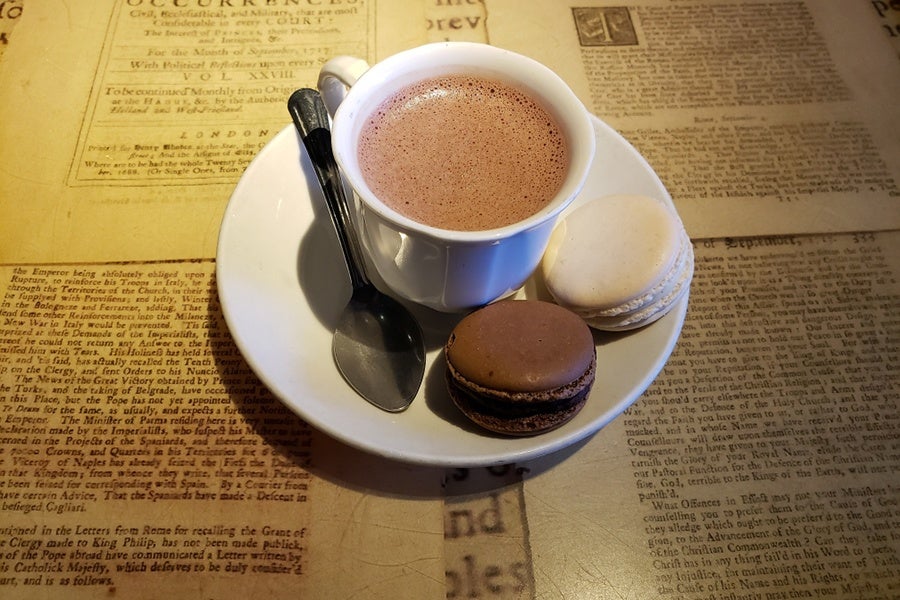 A mug of hot chocolate is accompanied by two macarons and sits on a table that looks like pages of a book.