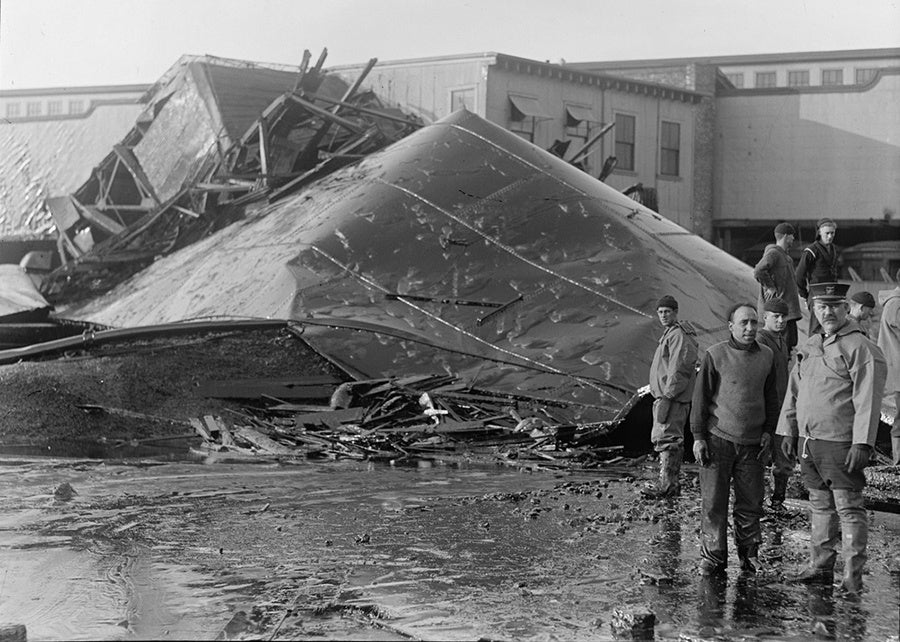 Historical photograph of the massive buckled steel wall of the collapsed molasses tank resting among wooden wreckage with investigators standing nearby.