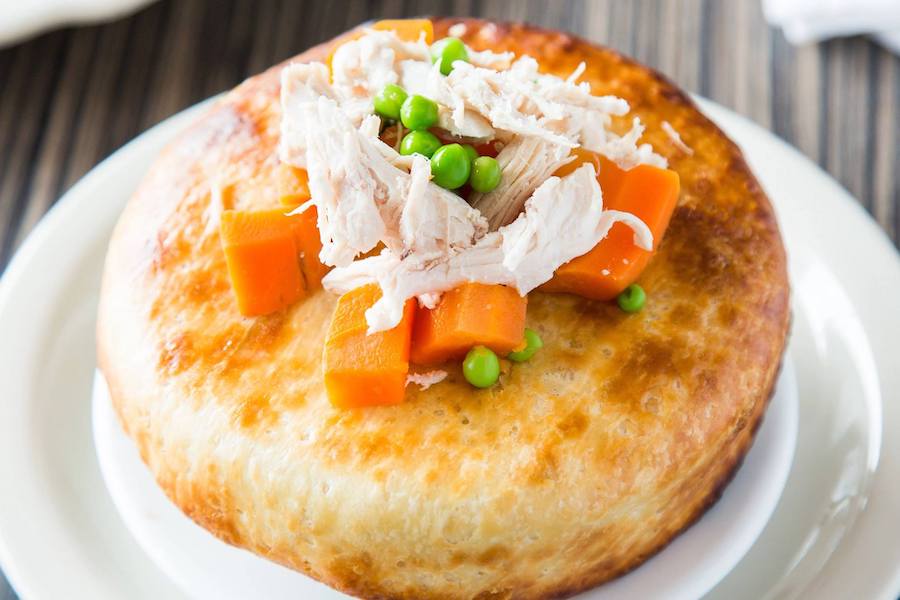 A round, golden brown pastry is topped with shredded chicken, carrot chunks, and peas.