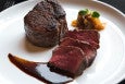 A plated dish featuring a thick, seared filet mignon steak, three slices of medium-rare beef, a dark glossy sauce, and a small garnish of caramelized onions topped with microgreens on a white plate.