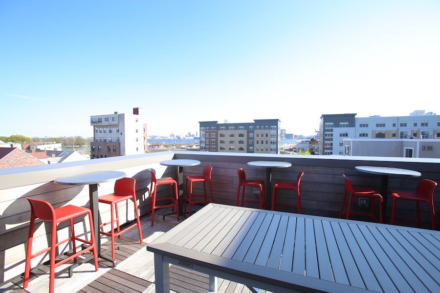 A minimalist city roof deck with light wooden accents and bright red high-top chairs.