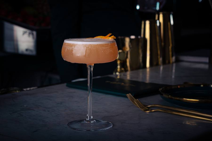 A pale orange cocktail in an elegant glass sits on a slate gray bar with gold flatware nearby.