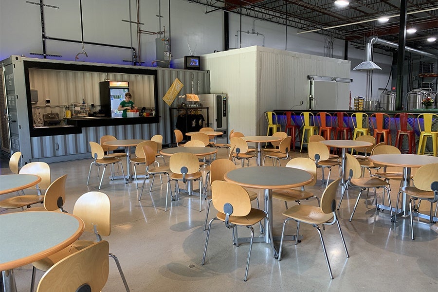 A brightly lit, empty taproom is filled with light wooden four-tops and colorful bar seating.