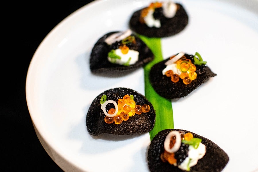 Five little black pancakes are lined up on a swoosh of green sauce on a white plate, each topped with orange salmon roe, a dollop of sour cream, and caviar.
