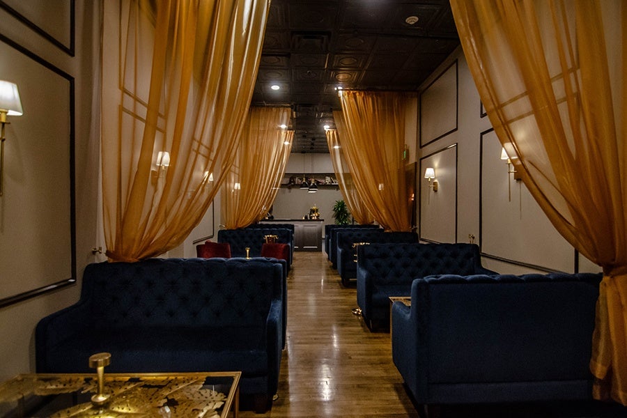 A dimly lit bar, long and narrow, features blue velvet couches and floor-to-ceiling sheer gold curtains near each.