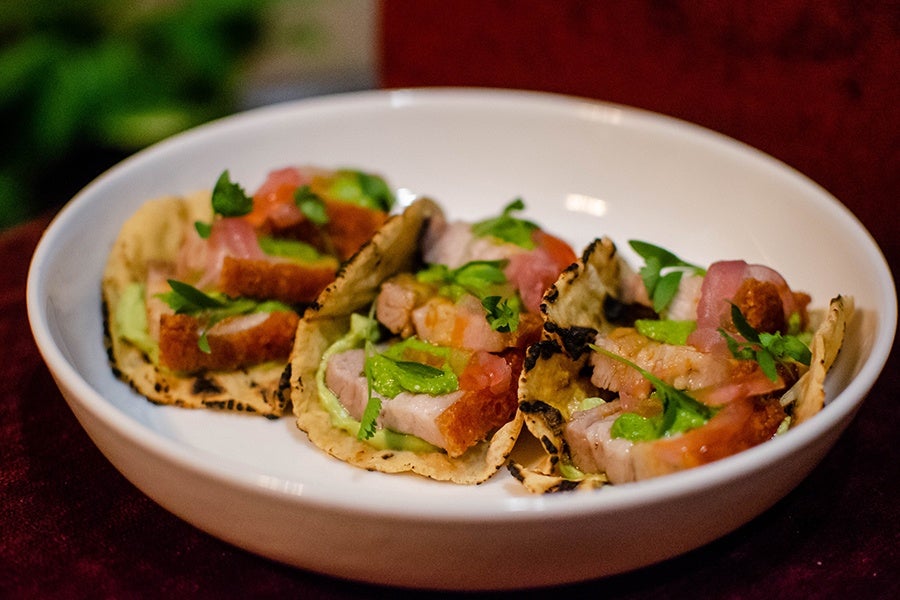 Three small tacos are stuffed with chunks of pork belly with crispy skin, purple rounds of pickled onions, and cilantro.
