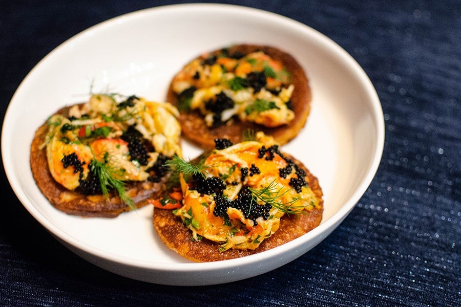 Three pancakes sit on a deep white plate, topped with pieces of lobster and tiny black dots of tobiko.