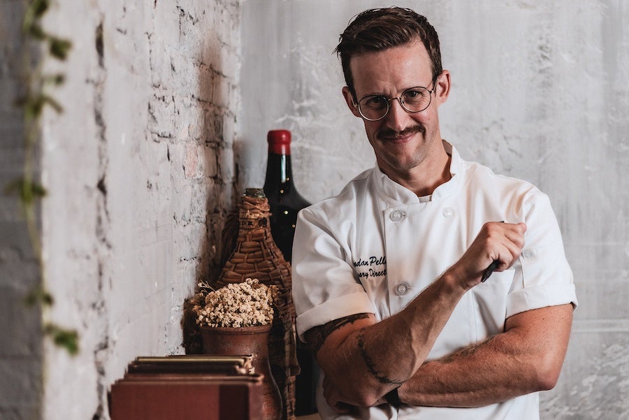 A man wearing chef's whites and glasses stares at the camera with a slight smile while leaning against a shelf of knick-knacks in a rustic restaurant space.