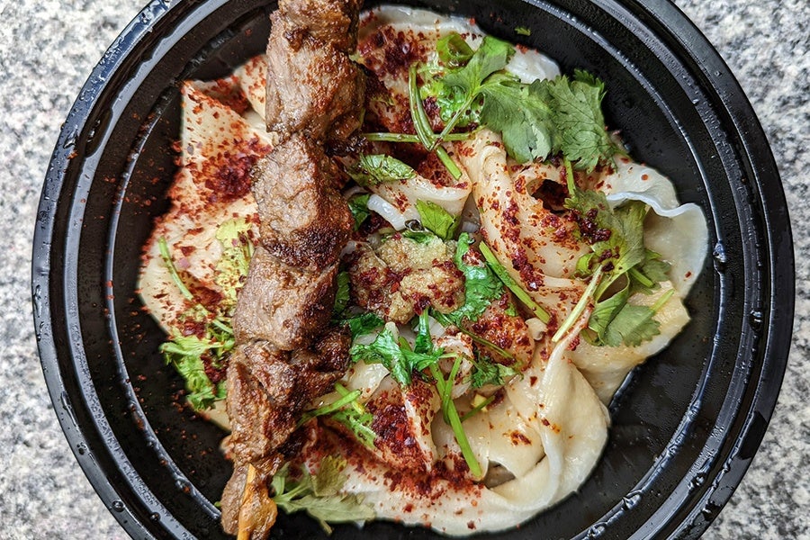 Takeout bowl of thick noodles dusted with chili powder and cilantro, garnished with a skewer of meat.