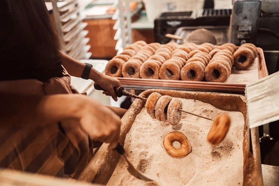 A person holds cider doughnuts on a rod, dipping them into a bucket of cinnamon sugar.