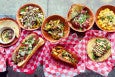 Overhead view of dishes of comfort food like mac and cheese, hot dogs, and tacos spread on red and white checkered paper.