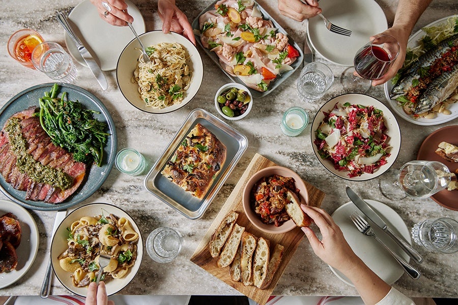Overhead view of a white marble table covered with Italian-inspired dishes, including bread and dips, steak, fish, pasta, and more.