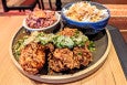 Two big pieces of crispy chicken are topped with a squiggle of a scallion sauce with small grapefruit wedges, and two bowls in the background hold red cabbage slaw and rice.