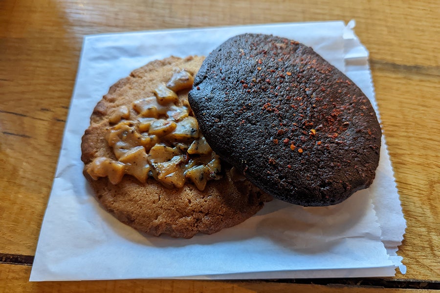 Two cookies, one topped with chunks of toffee and one dark chocolate with a dusting of red chili powder, sit on white paper.
