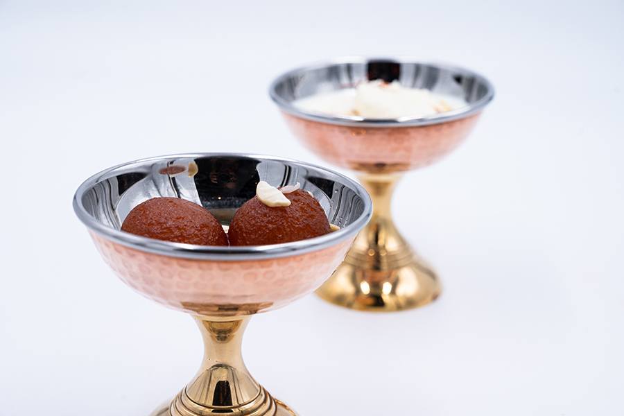 Two copper bowls are isolated on a white background, each holding an Indian dessert.