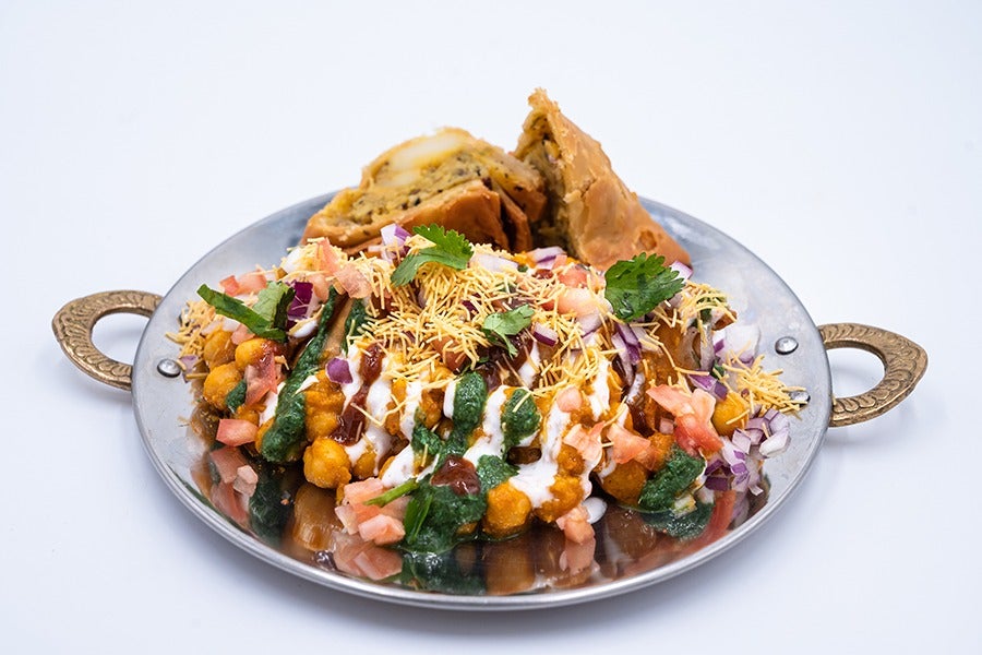 Indian chat—chickpeas, sauces, tomatoes, and other toppings—is displayed on a silver plate, with two halves of a samosa in the background.
