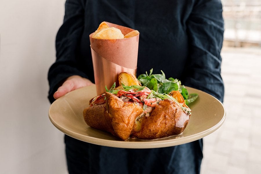 A restaurant server in black clothing holds out a plate with a lobster-stuffed popover and side of greens.