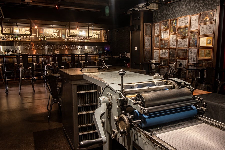 A bar with dim lighting features an old printing press and other printing press-related decor.