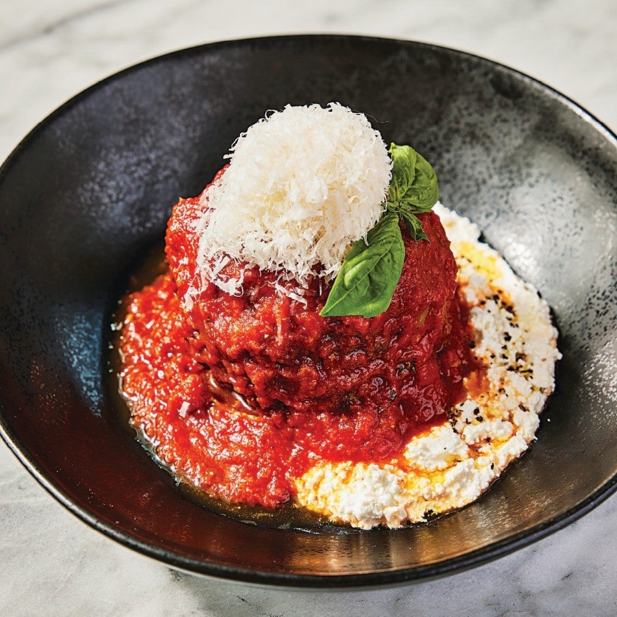 A giant, saucy meatball sits on a black plate, topped with a hefty amount of grated cheese and a basil leaf.