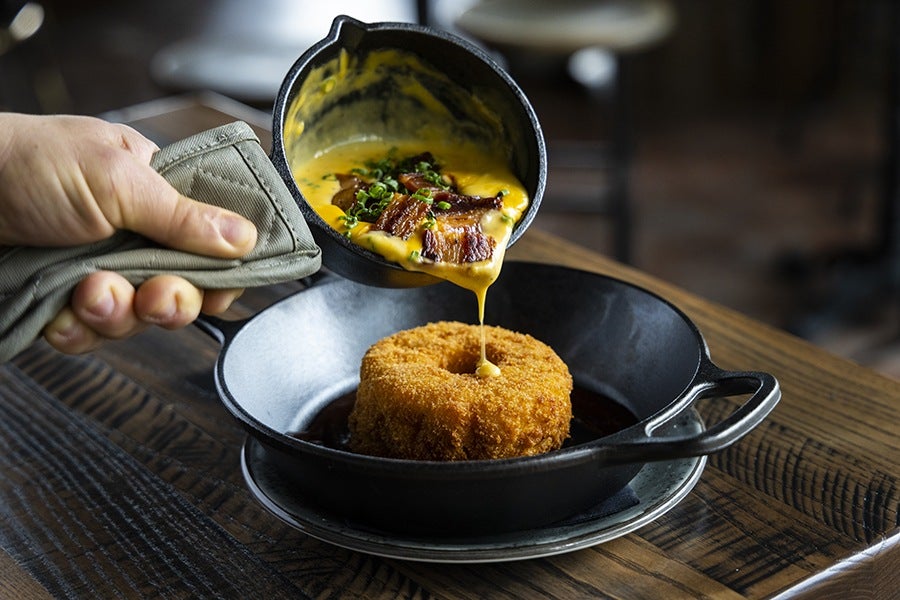 A hand pours a thick yellow-orange cheese sauce with pieces of bacon onto a breaded, fried doughnut.
