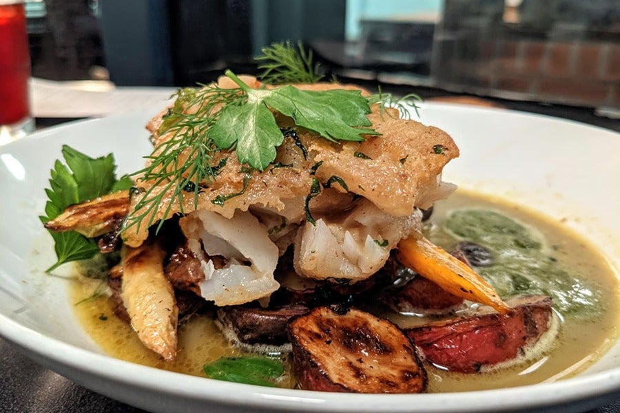 A flaky white fish sits atop rainbow carrots and potatoes in a thin light brown broth.