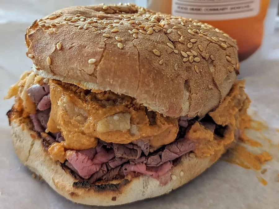 A sesame bun is piled high with rare, thinly sliced roast beef, topped with orange cheese sauce and crispy onions.