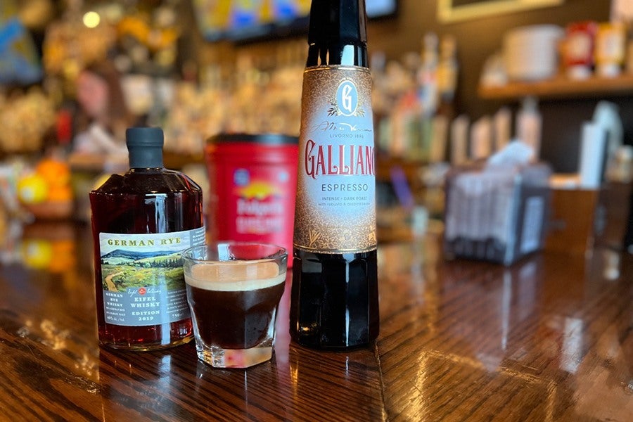 A dark brown cocktail is served in a short glass and displayed on a wooden bar, surrounded by bottles of its ingredients.