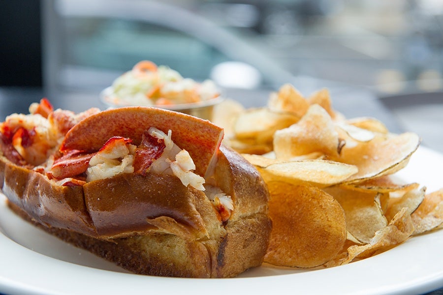 A grilled hot dog bun is stuffed full of lobster meat, with potato chips on the side.