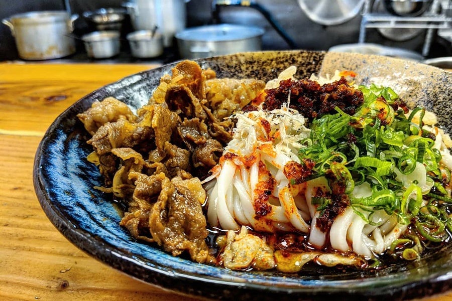 A bowl of thick udon noodles topped with chili paste, scallions, and thin slices of beef.