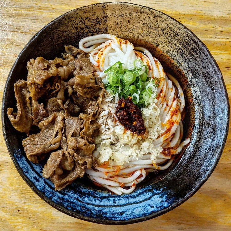 Overhead view of a bowl of thick udon noodles topped with chili paste, scallions, and thin slices of beef.