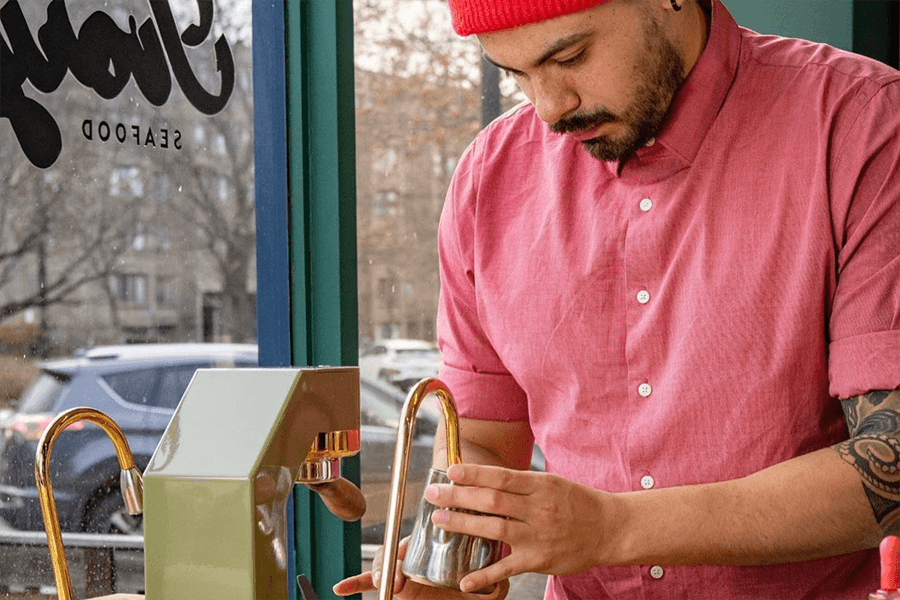 A man in a pink button down shirt and red knit cap makes coffee in a restaurant.