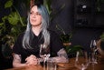 A woman with blue hair and a black button-down shirt sits at a table in a restaurant with lots of green plants in the background.