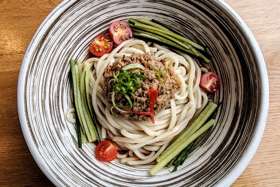 Overhead of a striking black and white striped bowl full of udon noodles, ground pork, cherry tomatoes, and cucumber sticks.