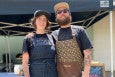 A man and a woman wearing chef's aprons and baseball caps smile, standing in front of an outdoor food pop-up setup.