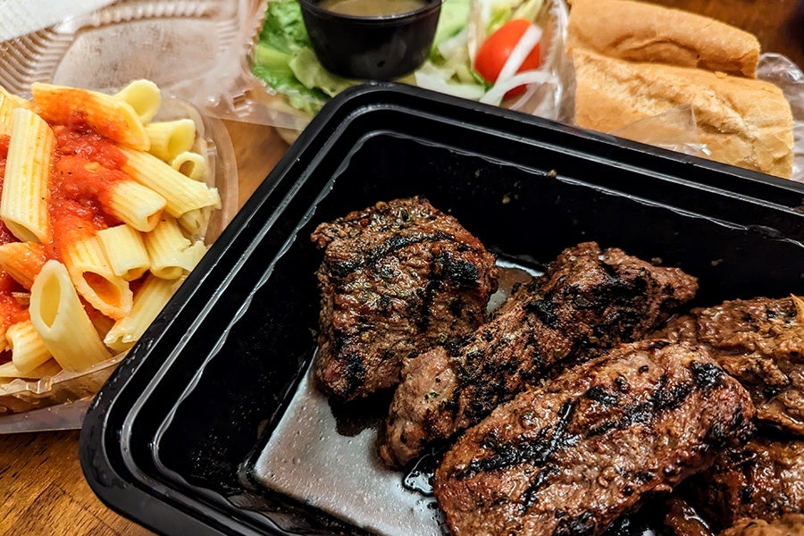 Large, grilled steak tips in a takeout container are accompanied by ziti with red sauce and a side salad.