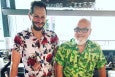 Two men stand smiling behind a bar, wearing Hawaiian shirts.