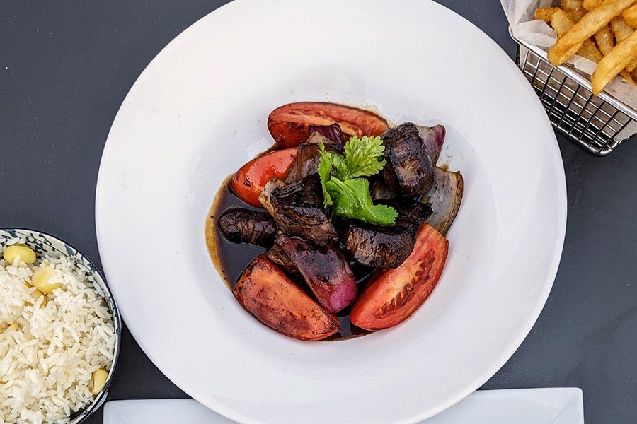 Overhead view of a dish of steak pieces in a dark marinade with tomatoes, onions, and sides of rice and fries.