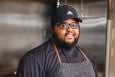 A bearded man in glasses and a baseball cap stands in a professional kitchen, wearing a denim apron and looking at the camera.