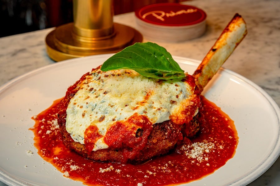 A thick-cut veal chop is breaded and fried, sitting in a pool of tomato sauce and covered with melted cheese and a basil leaf.
