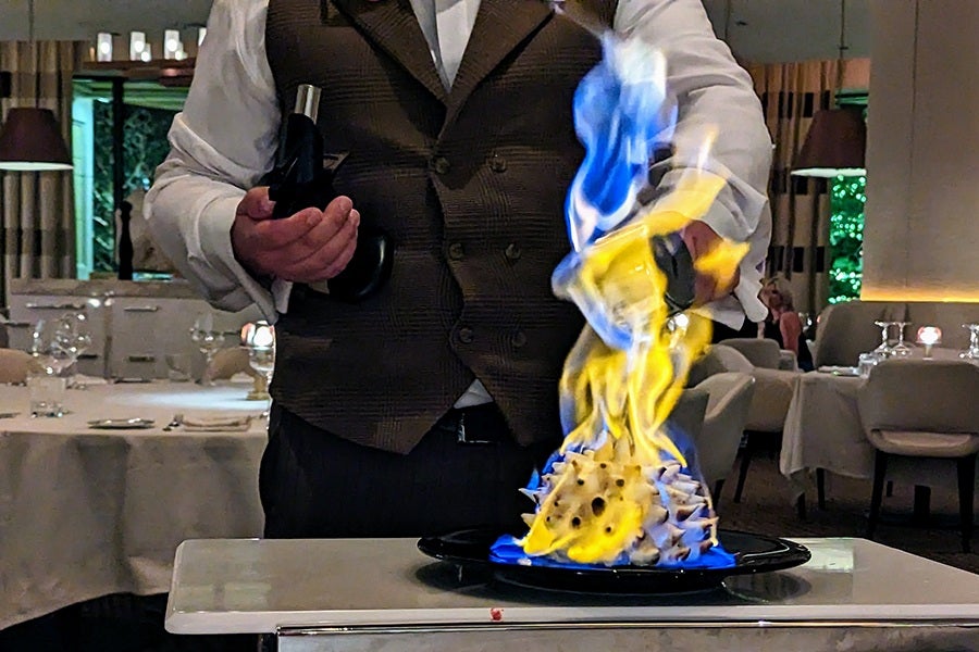 A fiery baked Alaska is prepared at a white-tablecloth restaurant by a server in a vest and button-down shirt.