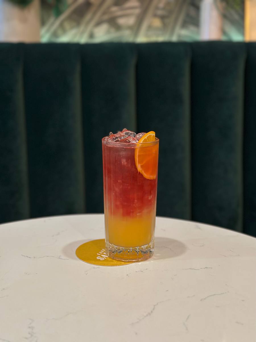 A mixed drink, served tall, has an orange layer, a pink layer, and an orange slice.