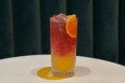 A mixed drink, served tall, has an orange layer, a pink layer, and an orange slice.