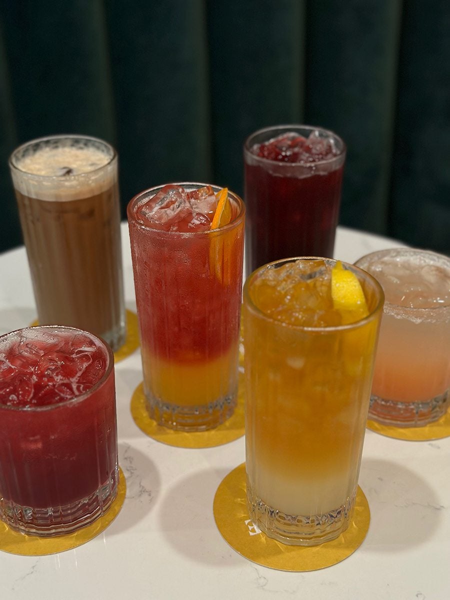 Five different mocktails sit on a white marble table with a green velvet couch in the background.