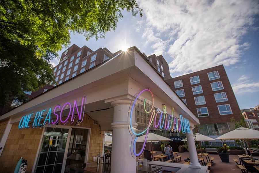 Picnic tables with umbrellas sit on a red brick courtyard next to a hotel, with an outdoor bar structure in the foreground with neon signage reading One Reason Garden Bar.