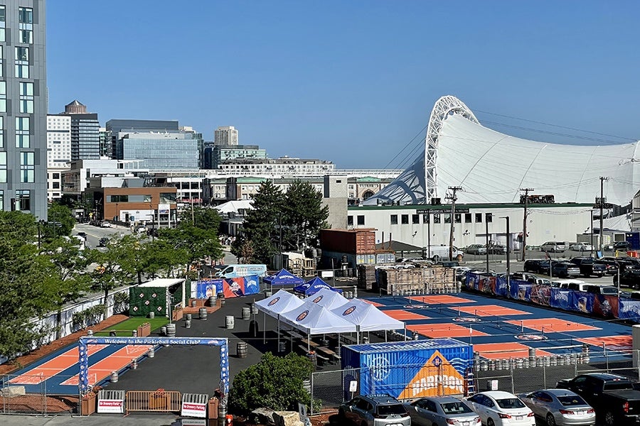In a break in the city skyline, there's an outdoor area with blue and orange pickleball and shuffleboard courts. Small tents to the side cover picnic tables.