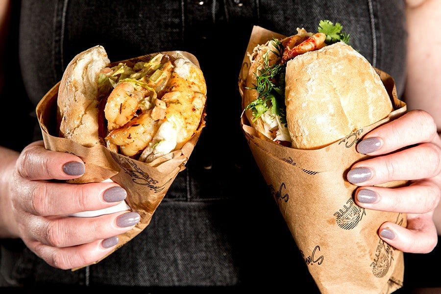 A hand holds two paper-wrapped sandwiches on baguettes.