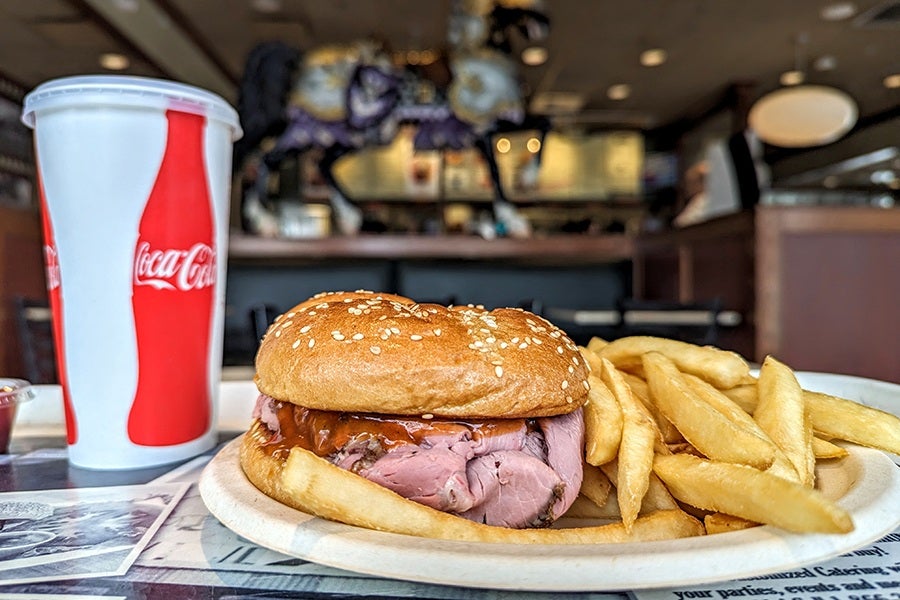 a roast beef sandwich and fries sit on a white paper plate inside a fast-food restaurant, accompanied by a cup of soda