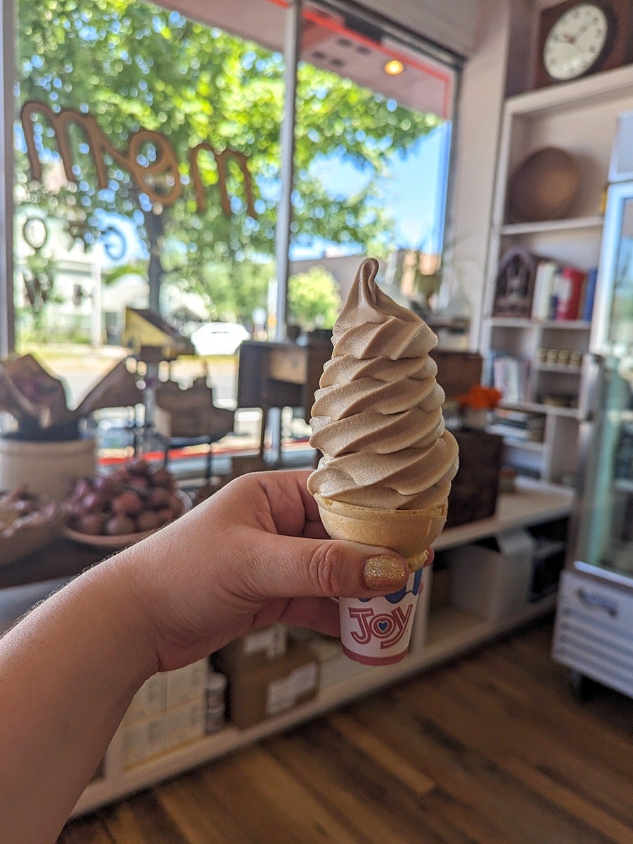 A hand holds a cone with a tall twist of beige and white soft-serve ice cream instead an old-timey grocery store.