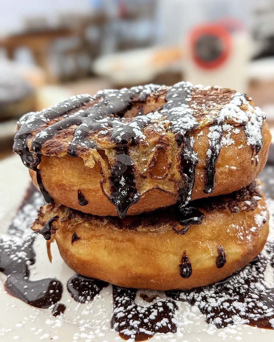 Two doughnuts are stacked on a plate, topped with chocolate sauce and powdered sugar.